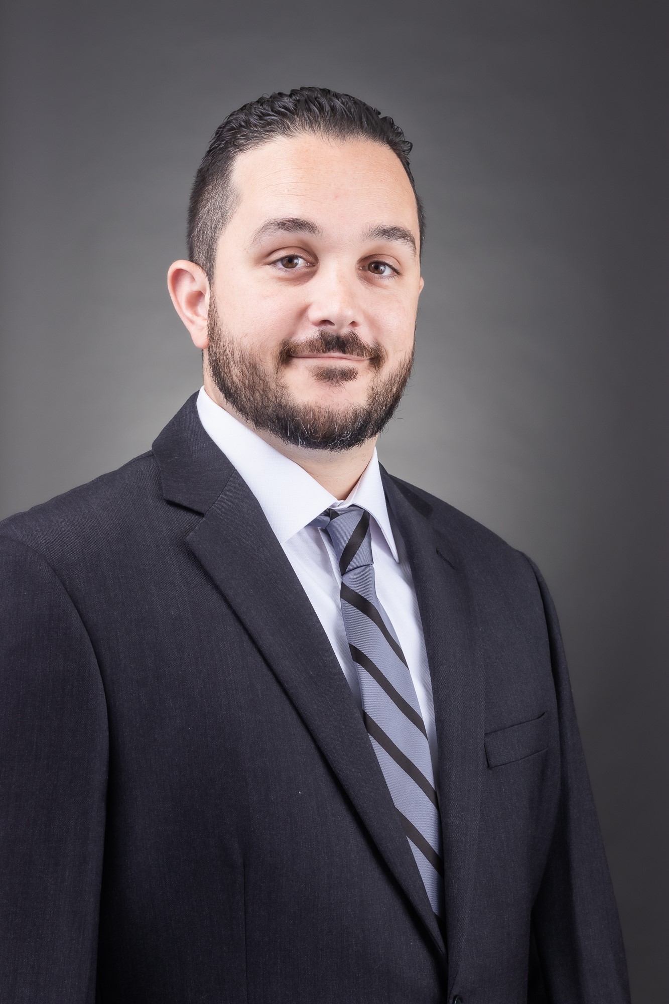 Professional headshot of Addison Verklas wearing a dark suit and tie with a neutral gray background